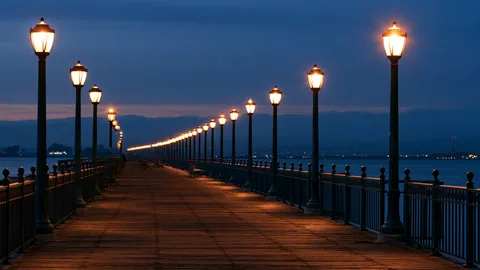 Symbolbild: Uferpromenade zur blauen Stunde mit einer langen Reihe warm leuchtender Laternen, die sich bis zum Horizont fortsetzt